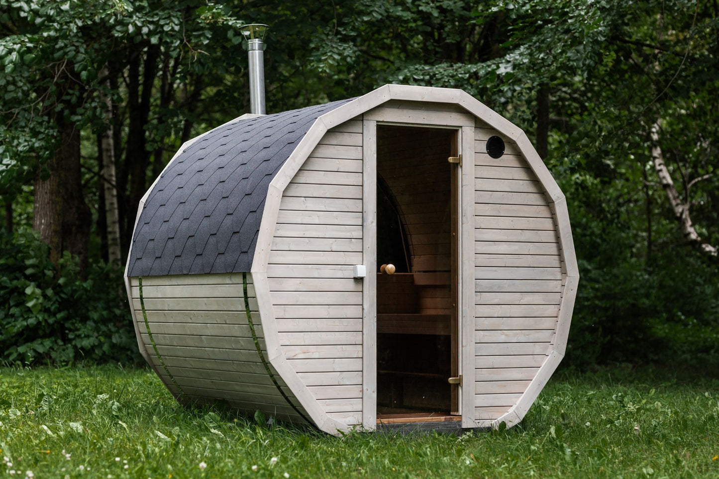 Runde Außensauna aus hochwertigem Fichten- oder Thermoholz, geeignet für 4–Personen. Mit Lindenholz-Bänken, Bitumenschindeln-Dach, Sicherheitsglas-Türen und Harvia-Beleuchtung.Außenansicht