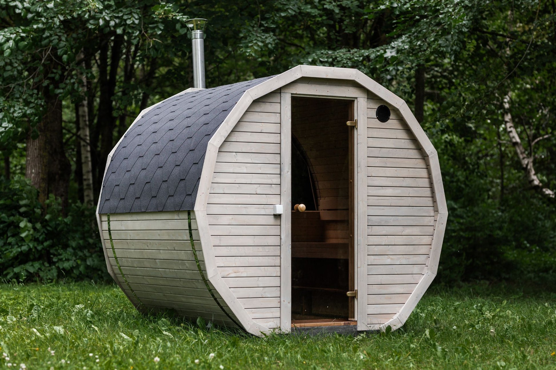 Runde Außensauna aus hochwertigem Fichten- oder Thermoholz, geeignet für 4–Personen. Mit Lindenholz-Bänken, Bitumenschindeln-Dach, Sicherheitsglas-Türen und Harvia-Beleuchtung.Außenansicht