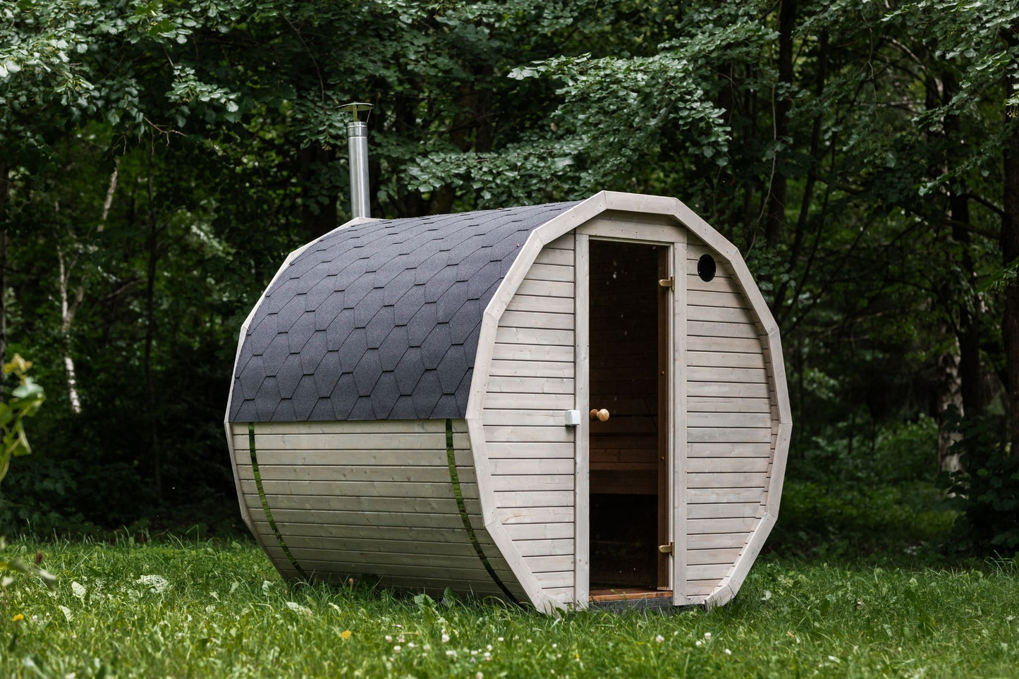 Runde Außensauna aus hochwertigem Fichten- oder Thermoholz, geeignet für 4–Personen. Mit Lindenholz-Bänken, Bitumenschindeln-Dach, Sicherheitsglas-Türen und Harvia-Beleuchtung.Außenansicht