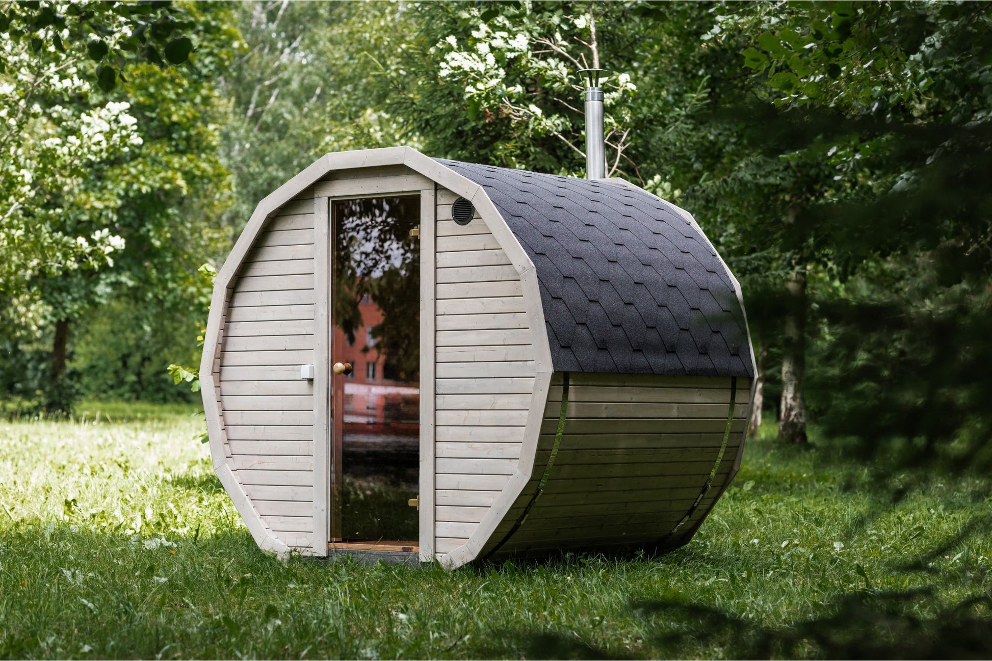 Runde Außensauna aus hochwertigem Fichten- oder Thermoholz, geeignet für 4–Personen. Mit Lindenholz-Bänken, Bitumenschindeln-Dach, Sicherheitsglas-Türen und Harvia-Beleuchtung.Außenansicht