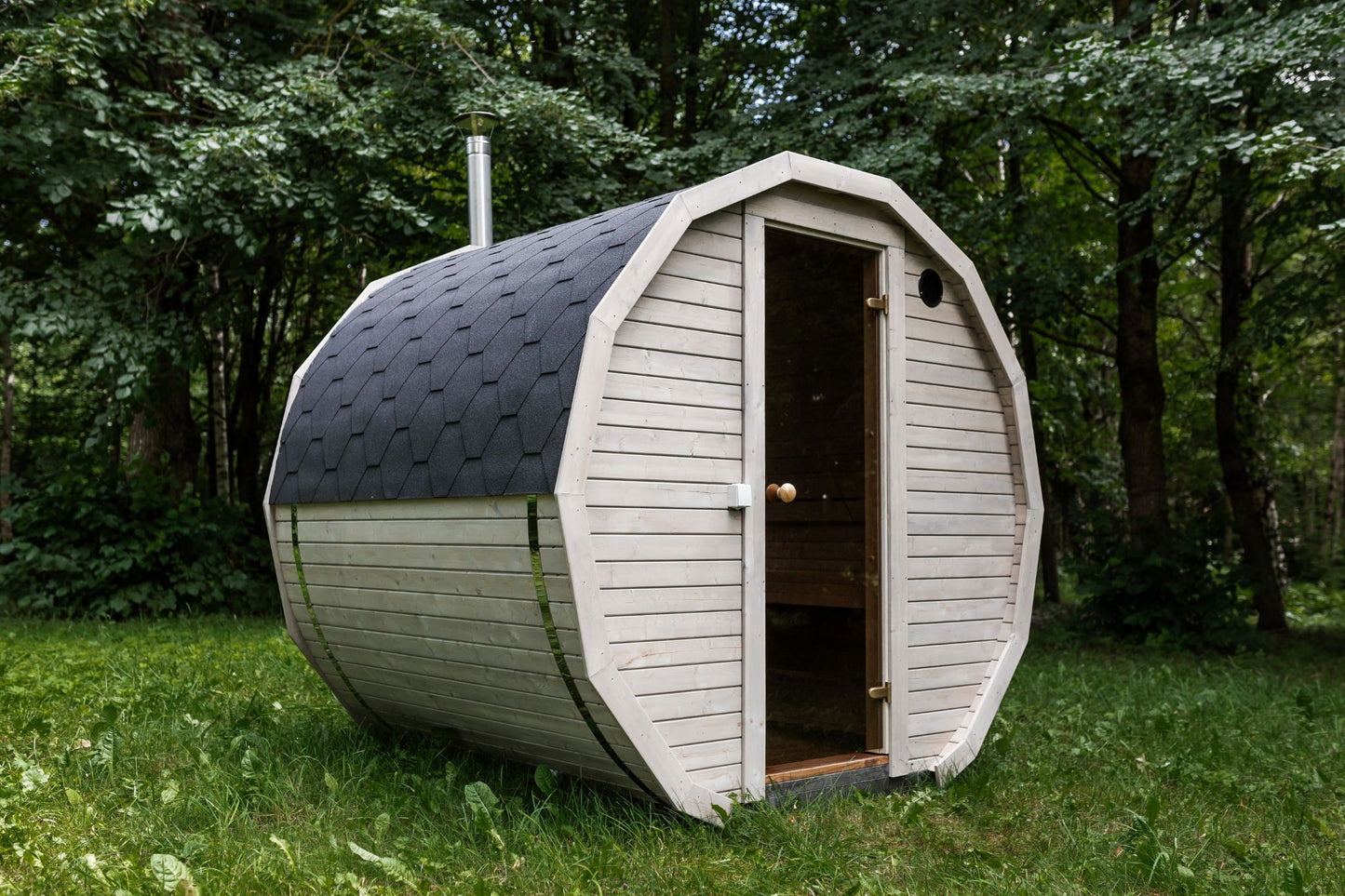Runde Außensauna aus hochwertigem Fichten- oder Thermoholz, geeignet für 4–Personen. Mit Lindenholz-Bänken, Bitumenschindeln-Dach, Sicherheitsglas-Türen und Harvia-Beleuchtung.Außenansicht
