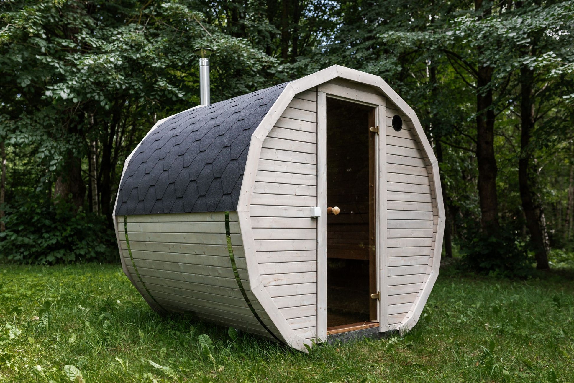 Runde Außensauna aus hochwertigem Fichten- oder Thermoholz, geeignet für 4–Personen. Mit Lindenholz-Bänken, Bitumenschindeln-Dach, Sicherheitsglas-Türen und Harvia-Beleuchtung.Außenansicht